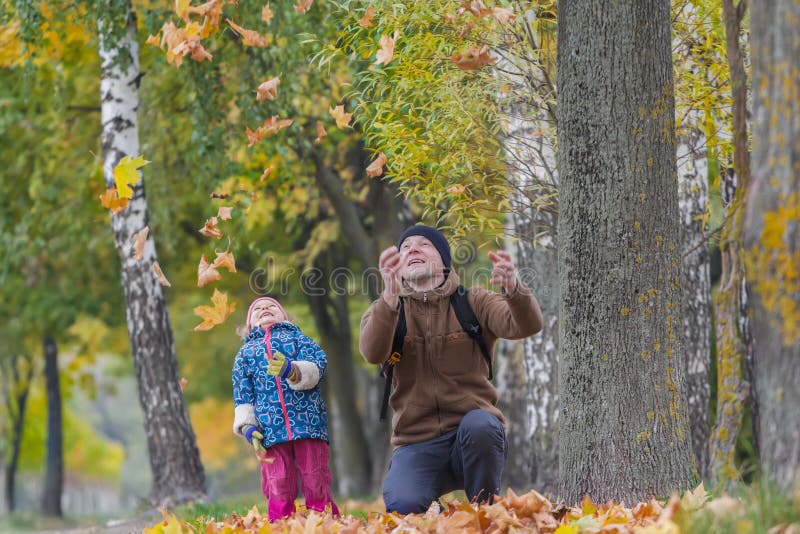 Père de sourire avec la fille riante jetant en l'air vers le haut des feuilles d'automne jaunes en parc dehors photographie stock