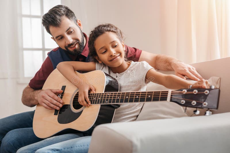 Père multi-ethnique et fille jouant la guitare sur le sofa à la maison photographie stock Père multi-ethnique et fille jouant la guitare sur le sofa à la maison photographie stock