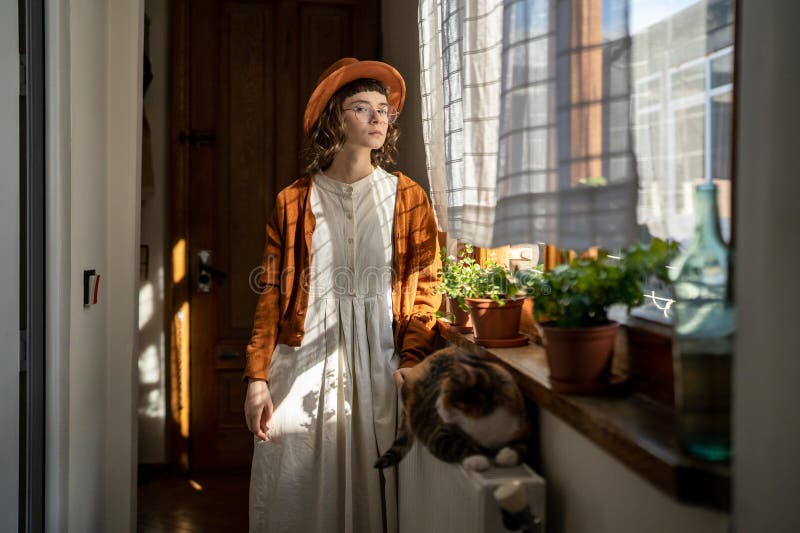 Pénitencière hipster fille propriétaire de animal debout près du chat moelleux couché sur la batterie de chauffage à la maison. Animal de compagnie à vie. pensive hipster girl portant chapeau et lunettes debout devant la fenêtre près du chaton moelleux allongé sur la batterie de chauffage. solitude et possession d'animaux de compagnie