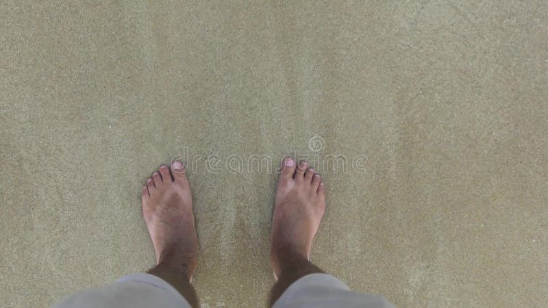 Pés masculinos no short que está na areia e nas ondas do ponto de vista da água do mar Pés masculinos em ondas do mar e na opiniã