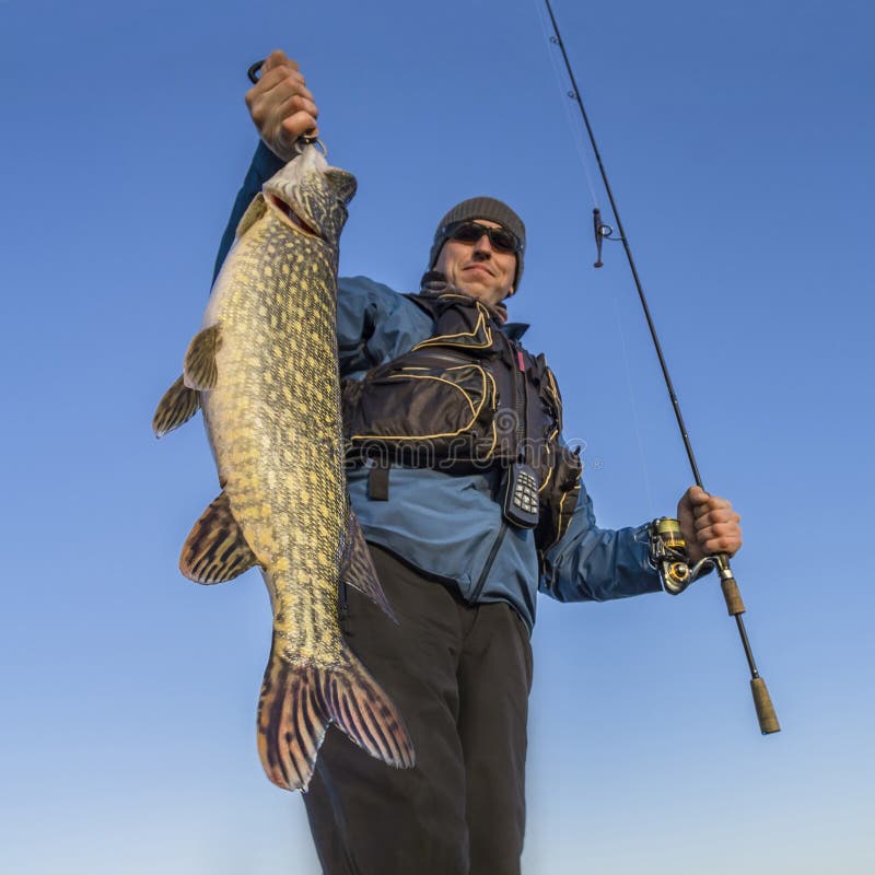 Happy fisherman holding big pike fish trophy and fishing rod on blue sky background. Happy fisherman holding big pike fish trophy and fishing rod on blue sky background