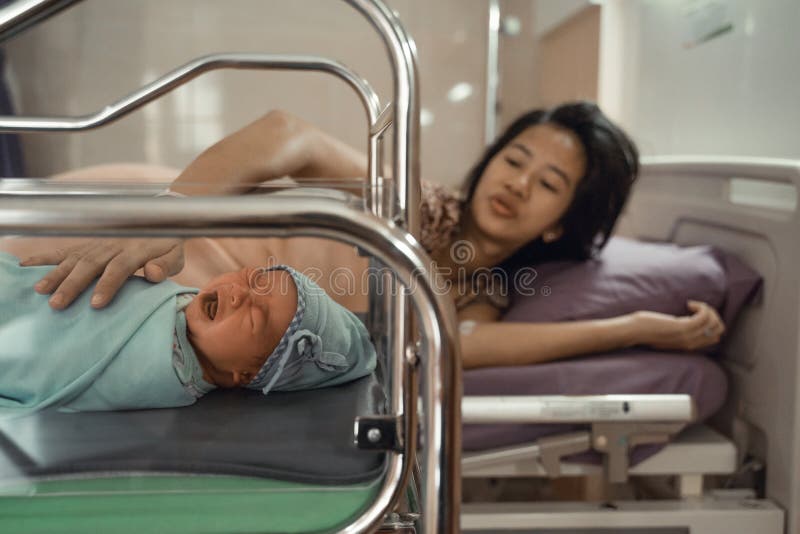 Crying asian newborn baby laying in crib with his mother lying on bed side in the hospital to comfort. Crying asian newborn baby laying in crib with his mother lying on bed side in the hospital to comfort