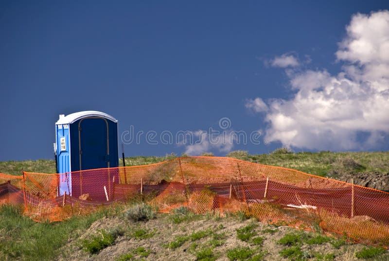 Blue portable toilet behind orange construction fence with blue sky and clouds. Blue portable toilet behind orange construction fence with blue sky and clouds