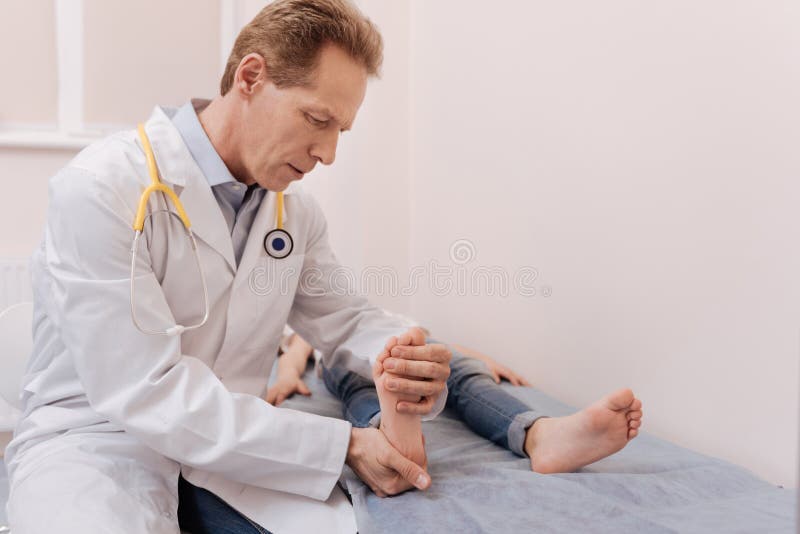 Careful moves. Prominent serious scrupulous doctor conducting a medical procedure and massaging girls feet while he lying on a bed. Careful moves. Prominent serious scrupulous doctor conducting a medical procedure and massaging girls feet while he lying on a bed