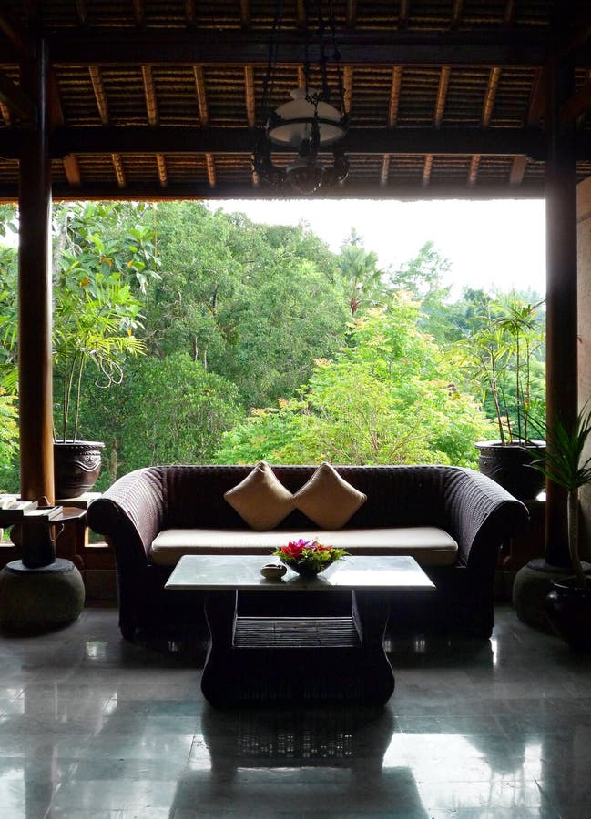 A colour photograph picture of a beautiful bali traditional style architecture with wood and bamboo furniture. Showing interior of a open style sitting room or patio balcony overlooking lush green valley forest. Room is furnished with coffee table, natural rattan cane sofa and cushions. Decorated with potted ornamental tropical plants and fresh flowers. Vertical color format photo. Nobody in picture. Location is upscale resort in Ubud, Bali, indonesia in southeast asia. Travels photography to exotic destinations with local architectural and deco interest. A colour photograph picture of a beautiful bali traditional style architecture with wood and bamboo furniture. Showing interior of a open style sitting room or patio balcony overlooking lush green valley forest. Room is furnished with coffee table, natural rattan cane sofa and cushions. Decorated with potted ornamental tropical plants and fresh flowers. Vertical color format photo. Nobody in picture. Location is upscale resort in Ubud, Bali, indonesia in southeast asia. Travels photography to exotic destinations with local architectural and deco interest.