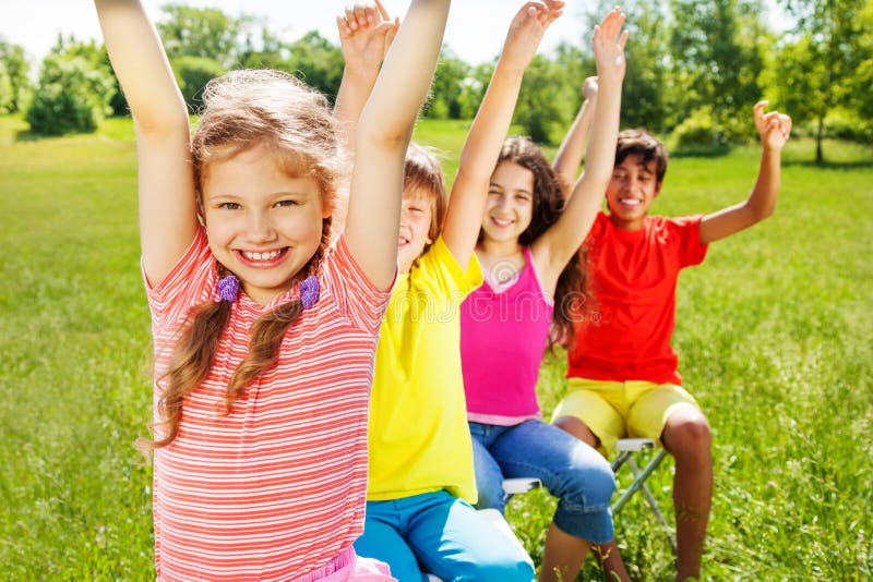 Four kids sitting in row on chairs with hands up. Four kids sitting in row on chairs with hands up.