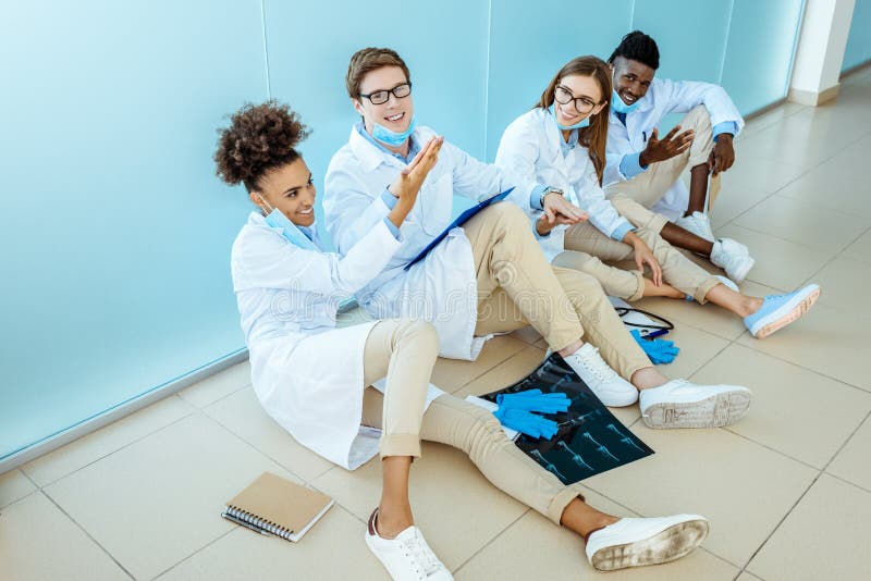 Four smiling young medical interns in white robes sitting on a floor in hospital high-fiving and celebrating success. Four smiling young medical interns in white robes sitting on a floor in hospital high-fiving and celebrating success