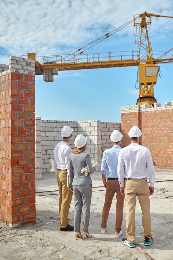 Quattro persone con caschi di protezione che ispezionano l'area dell'edificio. Retrovisore di un supervisore con disegni tecnici e costruttori in piedi sul cantiere