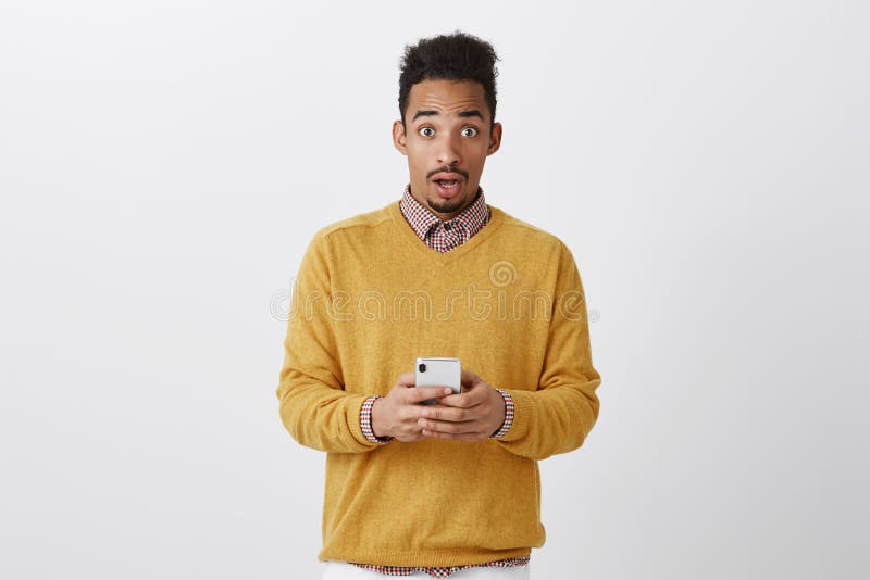 Someone tried to hack his phone. Shocked handsome boyfriend with afro hairstyle in trendy clothes holding smartphone, staring at camera with amazed expression, being surprised over gray wall. Someone tried to hack his phone. Shocked handsome boyfriend with afro hairstyle in trendy clothes holding smartphone, staring at camera with amazed expression, being surprised over gray wall.