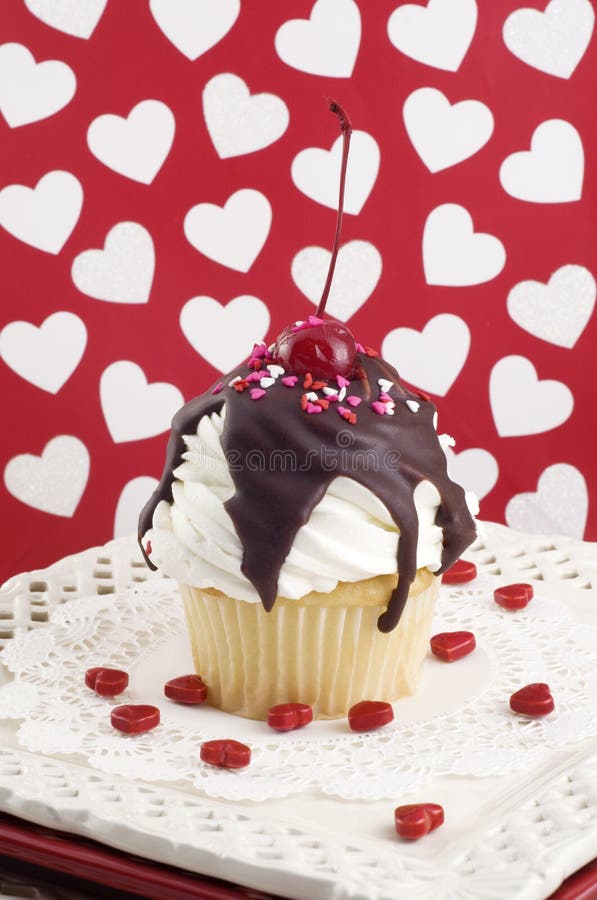 A Valentine sundae cupcake with white frosting, fudge, topped with a cherry and sprinkles, heart background, selective focus, vertical. A Valentine sundae cupcake with white frosting, fudge, topped with a cherry and sprinkles, heart background, selective focus, vertical