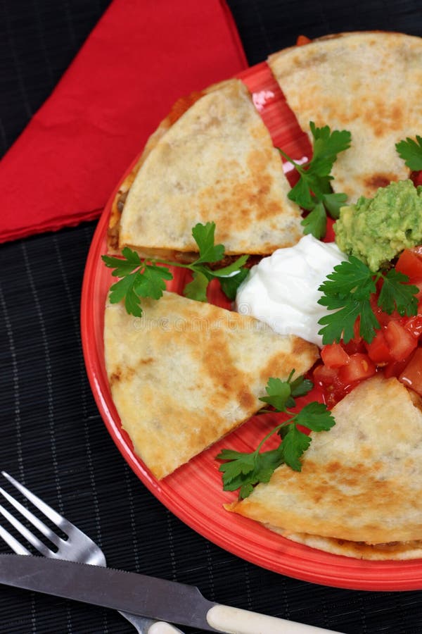 Mexican quesadillas served with tomato salsa, guacamole and sour cream. Served on a dark pink plate and garnished with sprigs of parsley. On a black background with a knife and fork and a red napkin.