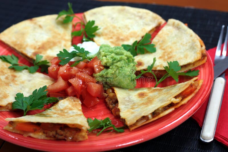 A plate of quesadillas filled with meat sauce and cheese served with tomato salsa, guacamole and sour cream. Served on a pink plate and garnished with fresh parsley. On a black background.