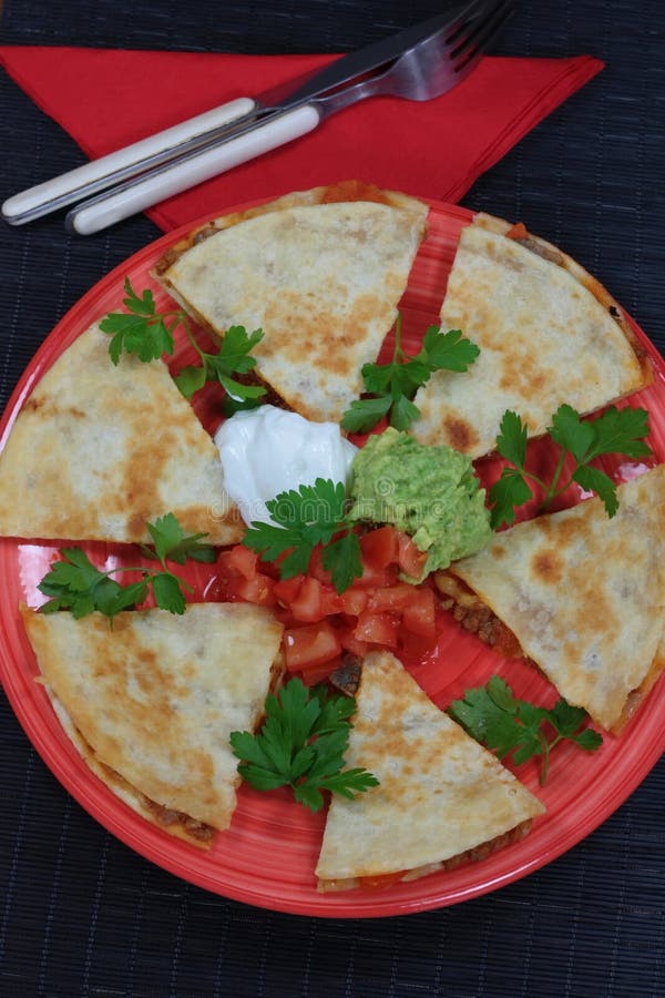 A plate of delicious Mexican Quesadillas filled with meat sauce, tomtoes and cheese and served with a trio of spicy tomato salsa, sour cream and fresh guacamole. Viewed from above and on a black background. A plate of delicious Mexican Quesadillas filled with meat sauce, tomtoes and cheese and served with a trio of spicy tomato salsa, sour cream and fresh guacamole. Viewed from above and on a black background.