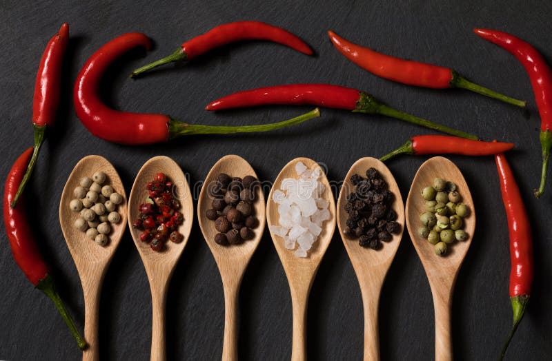 Different kind of pepper and salt in wooden spoons on a black slate background