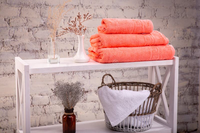 Rack with a stack of three peach color towels and baskets with clean white towels and toilet decor near a brick wall. Shelf with