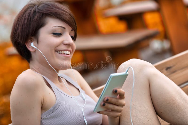 Blissful young woman smiling to herself as she listens to music while relaxing outdoors on a park bench in a close up view. Blissful young woman smiling to herself as she listens to music while relaxing outdoors on a park bench in a close up view