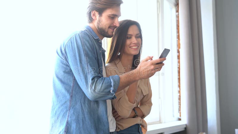 Radosna para patrząca na telefon komórkowy w domu. uśmiechnięty mąż przytulający żonę przy oknie
