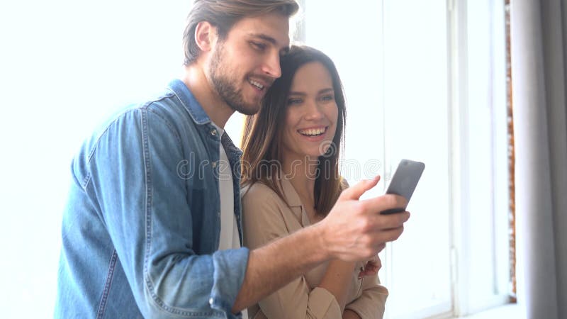 Radosna para patrząca na telefon komórkowy w domu. uśmiechnięty mąż przytulający żonę przy oknie