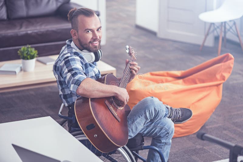 Radosny mężczyzna na wózku inwalidzkim bawić się gitarę w rozsądnym studiu nagrań obraz royalty free Radosny mężczyzna na wózku inwalidzkim bawić się gitarę w rozsądnym studiu nagrań obraz royalty free