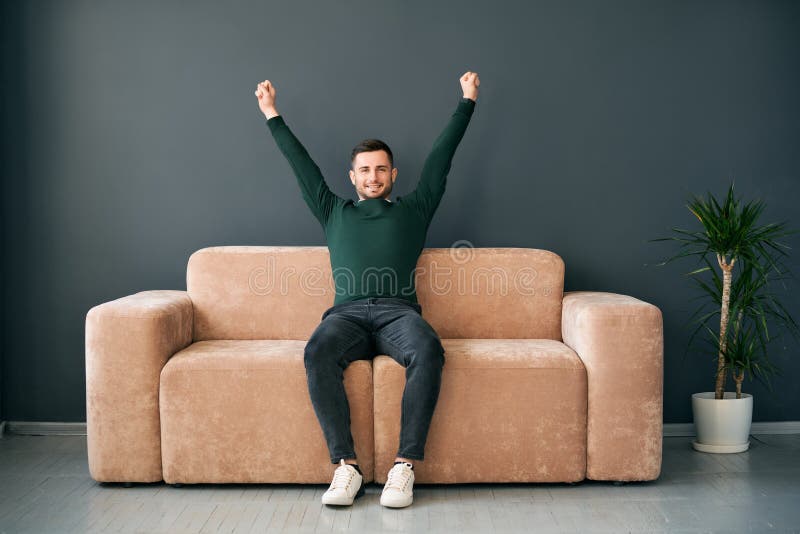 Radosny młody mężczyzna z ramionami wzniesionymi, uczcząc sukces siedzący na sofie we współczesnym mieszkaniu