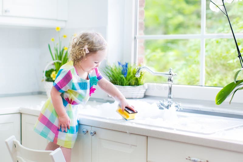 Cute curly toddler girl in a colorful dress washing dishes, cleaning with a sponge and playing with foam in the sink in a beautiful sunny white kitchen with a garden view window in a modern home. Cute curly toddler girl in a colorful dress washing dishes, cleaning with a sponge and playing with foam in the sink in a beautiful sunny white kitchen with a garden view window in a modern home