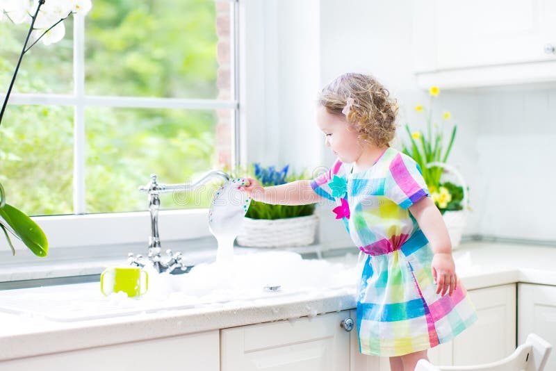 Cute curly toddler girl in a colorful dress washing dishes, cleaning with a sponge and playing with foam in the sink in a beautiful sunny white kitchen with a garden view window in a modern home. Cute curly toddler girl in a colorful dress washing dishes, cleaning with a sponge and playing with foam in the sink in a beautiful sunny white kitchen with a garden view window in a modern home