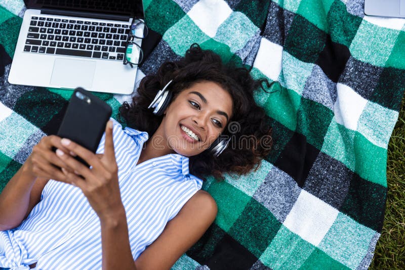 Ragazza africana che utilizza aria aperta del telefono cellulare nella musica d'ascolto del parco con le cuffie fotografie stock libere da diritti Ragazza africana che utilizza aria aperta del telefono cellulare nella musica d'ascolto del parco con le cuffie fotografie stock libere da diritti