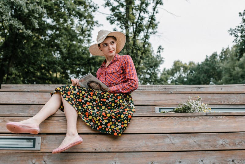 Ragazza alla moda che legge un libro Una bella donna con un cappello che si siede sulle scale di un albero Uno studente legge un immagine stock libera da diritti Ragazza alla moda che legge un libro Una bella donna con un cappello che si siede sulle scale di un albero Uno studente legge un immagine stock libera da diritti
