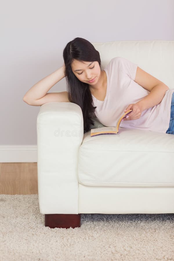 Ragazza asiatica contenta che si trova sul libro di lettura del sofà fotografia stock