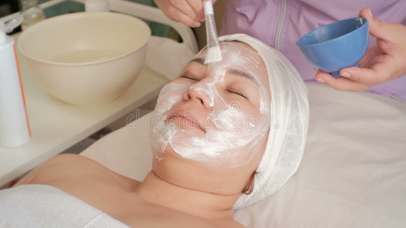 Ragazza asiatica in un salone di bellezza Video accelerato L'estetista applica una crema bianca di massaggio al fronte di una don