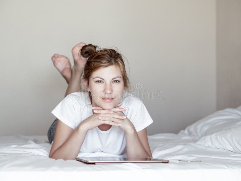 Ragazza castana attraente che si trova sul letto accanto alla compressa ed al telefono e che sorride alla macchina fotografica in fotografia stock libera da diritti Ragazza castana attraente che si trova sul letto accanto alla compressa ed al telefono e che sorride alla macchina fotografica in fotografia stock libera da diritti
