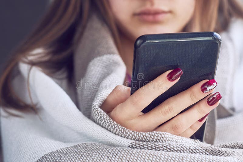 Ragazza che si siede sul davanzale della finestra con lo smartphone nero in mani avvolte in una coperta calda della lana immagine stock Ragazza che si siede sul davanzale della finestra con lo smartphone nero in mani avvolte in una coperta calda della lana immagine stock