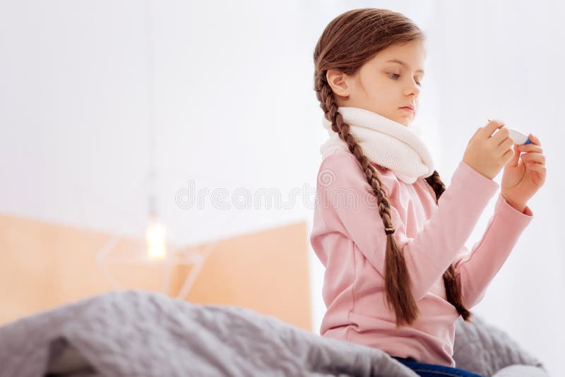 Ragazza curiosa che misura la temperatura mentre sedendosi sul letto fotografia stock Ragazza curiosa che misura la temperatura mentre sedendosi sul letto fotografia stock