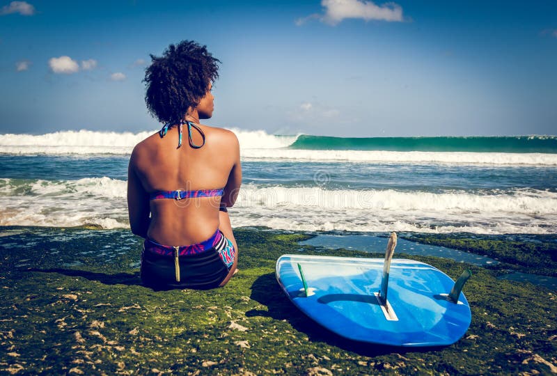 Ragazza del surfista con l'acconciatura di afro che si siede accanto al surf blu sulla barriera corallina verde fotografia stock Ragazza del surfista con l'acconciatura di afro che si siede accanto al surf blu sulla barriera corallina verde fotografia stock