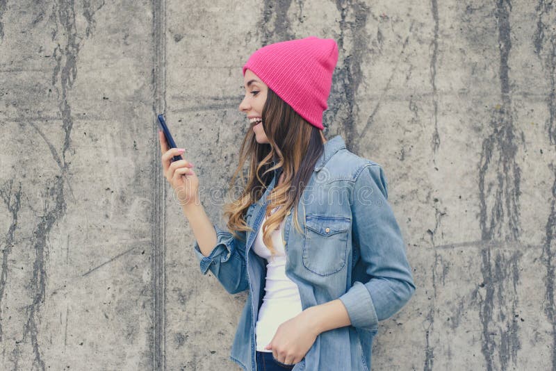 Ragazza emozionante felice di risata in abbigliamento dei jeans e cappello rosa facendo uso dello smartphone e macchina fotografi immagine stock libera da diritti Ragazza emozionante felice di risata in abbigliamento dei jeans e cappello rosa facendo uso dello smartphone e macchina fotografi immagine stock libera da diritti