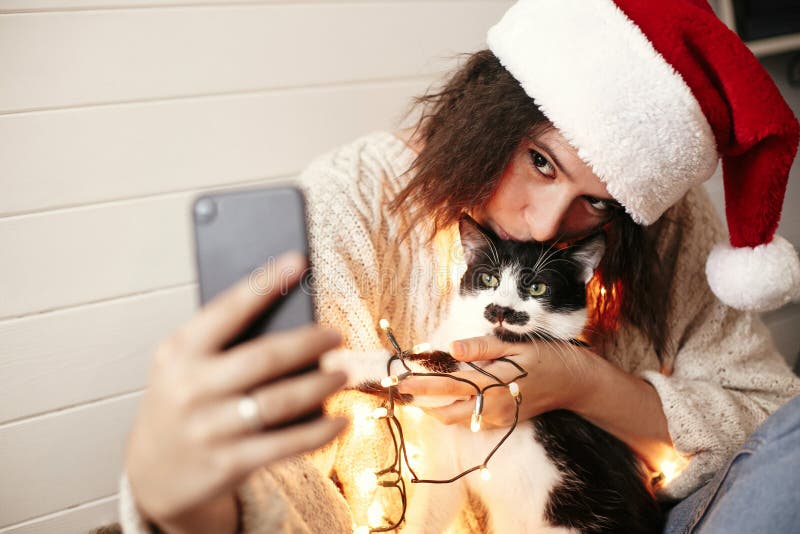 Ragazza felice alla moda in cappello di Santa che prende selfie con il gatto sveglio alle luci di natale su fondo dei regali Giov fotografie stock Ragazza felice alla moda in cappello di Santa che prende selfie con il gatto sveglio alle luci di natale su fondo dei regali Giov fotografie stock