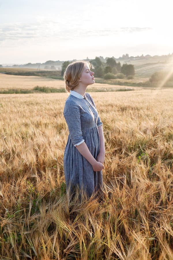 Ragazza felice piacevole in vestito all'aperto antiquato blu nel campo dorato delle orecchie ad alba Alba nebbiosa nel fondo fotografie stock Ragazza felice piacevole in vestito all'aperto antiquato blu nel campo dorato delle orecchie ad alba Alba nebbiosa nel fondo fotografie stock