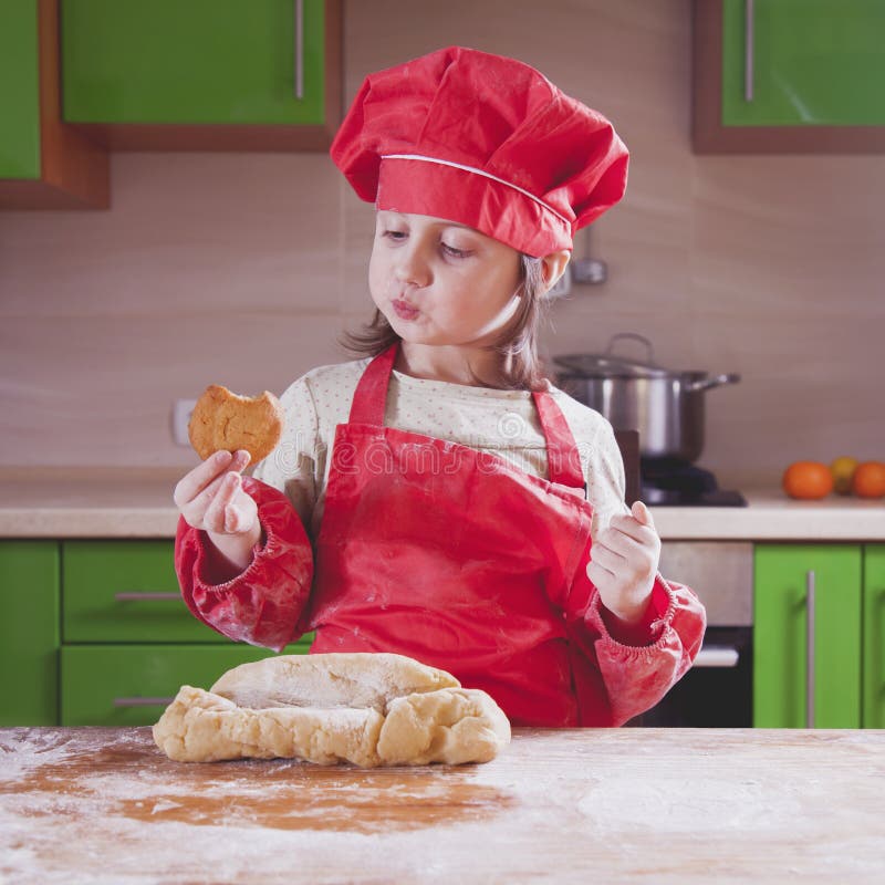 Ragazza graziosa del bambino piccolo con il dolce e la degustazione della tenuta del cappello del cuoco unico Alimento, processo immagini stock libere da diritti Ragazza graziosa del bambino piccolo con il dolce e la degustazione della tenuta del cappello del cuoco unico Alimento, processo immagini stock libere da diritti