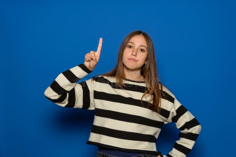 Ragazza ispanica di circa 15 anni con un maglione a strisce su sfondo blu puntando il dito verso l'alto e apparendo ispirata immagine stock