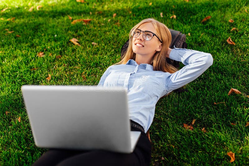 Ragazza pensierosa in vetri che si trovano sull'erba nel parco con un computer portatile, sogni di qualcosa immagine stock libera da diritti Ragazza pensierosa in vetri che si trovano sull'erba nel parco con un computer portatile, sogni di qualcosa immagine stock libera da diritti