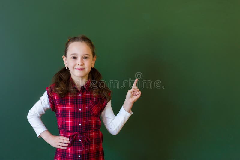 Ragazza prescolare della scolara felice nella condizione del vestito dal plaid nella classe vicino ad una lavagna verde Concetto fotografia stock libera da diritti Ragazza prescolare della scolara felice nella condizione del vestito dal plaid nella classe vicino ad una lavagna verde Concetto fotografia stock libera da diritti
