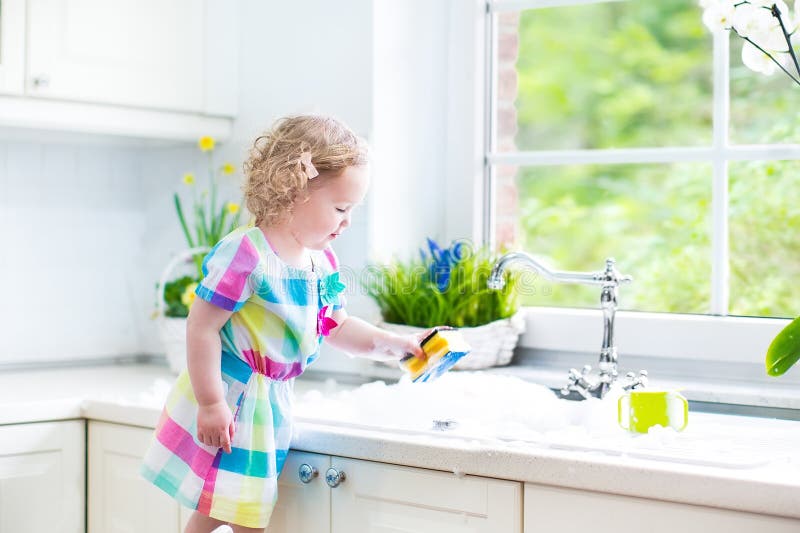 Cute curly toddler girl in a colorful dress washing dishes, cleaning with a sponge and playing with foam in the sink in a beautiful sunny white kitchen with a garden view window in a modern home. Cute curly toddler girl in a colorful dress washing dishes, cleaning with a sponge and playing with foam in the sink in a beautiful sunny white kitchen with a garden view window in a modern home