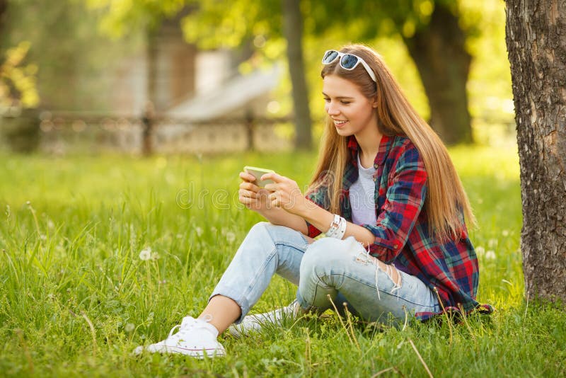 Ragazza sorridente attraente che scrive sul telefono cellulare nel parco della città di estate Donna felice moderna con uno smart fotografia stock libera da diritti Ragazza sorridente attraente che scrive sul telefono cellulare nel parco della città di estate Donna felice moderna con uno smart fotografia stock libera da diritti