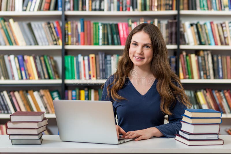 Ragazza sorridente che si siede ad uno scrittorio nella biblioteca che lavora con immagini stock libere da diritti Ragazza sorridente che si siede ad uno scrittorio nella biblioteca che lavora con immagini stock libere da diritti