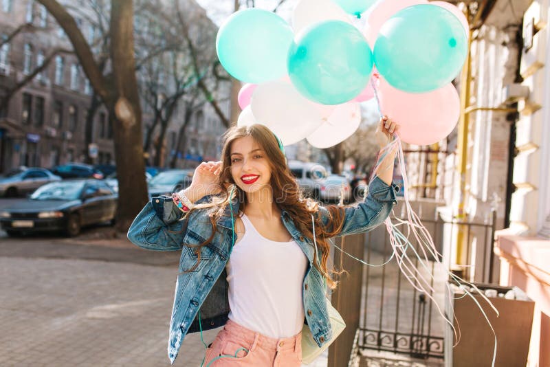 Ragazza sorridente incantante in rivestimento del denim che posa con il mazzo di palloni variopinti sui precedenti della città Ri fotografia stock libera da diritti Ragazza sorridente incantante in rivestimento del denim che posa con il mazzo di palloni variopinti sui precedenti della città Ri fotografia stock libera da diritti