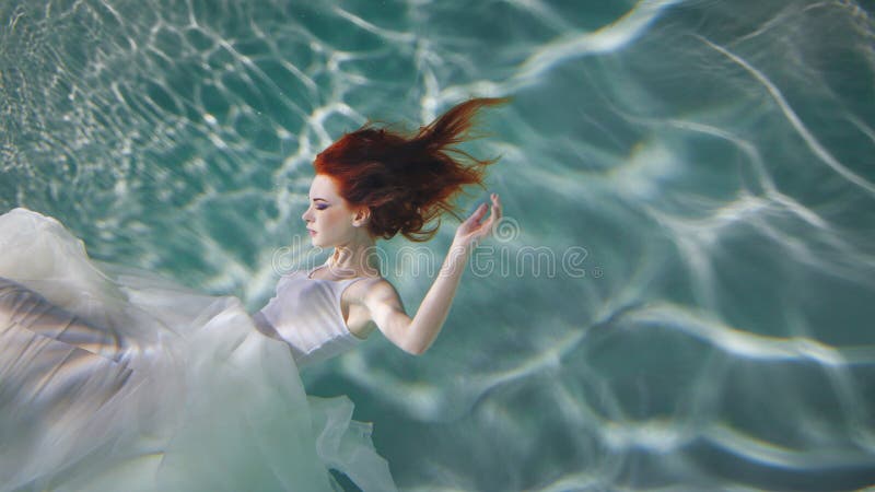Ragazza subacquea Bella donna dai capelli rossi in un vestito bianco, nuotante sotto l'acqua fotografia stock libera da diritti Ragazza subacquea Bella donna dai capelli rossi in un vestito bianco, nuotante sotto l'acqua fotografia stock libera da diritti