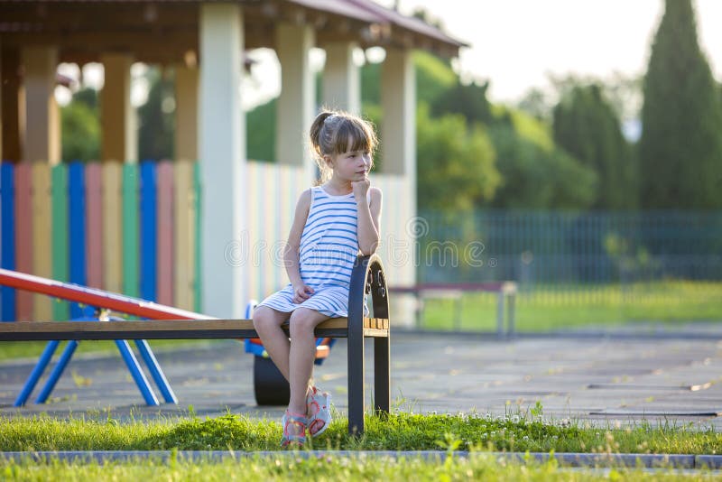 Ragazza sveglia in breve vestito che si siede da solo all'aperto sul banco del campo da giuoco il giorno di estate soleggiato fotografia stock Ragazza sveglia in breve vestito che si siede da solo all'aperto sul banco del campo da giuoco il giorno di estate soleggiato fotografia stock