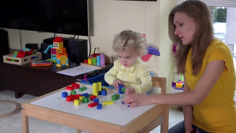 Ragazza sveglia con la madre che gioca i mattoni di legno variopinti vicino alla piccola tavola a casa archivi video