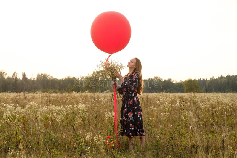 Ragazza in un bello vestito con i fiori con un pallone rosso su un guinzaglio nel campo al tramonto fotografia stock Ragazza in un bello vestito con i fiori con un pallone rosso su un guinzaglio nel campo al tramonto fotografia stock