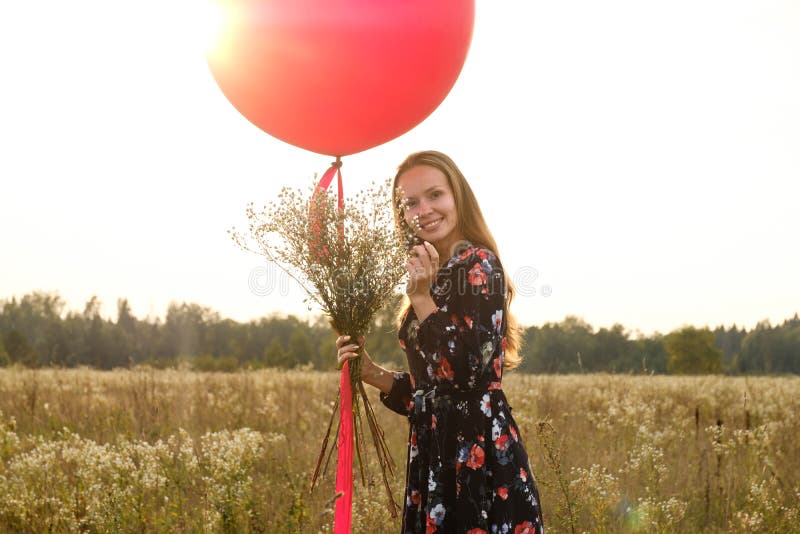 Ragazza in un bello vestito con i fiori con un pallone rosso su un guinzaglio nel campo al tramonto fotografie stock Ragazza in un bello vestito con i fiori con un pallone rosso su un guinzaglio nel campo al tramonto fotografie stock